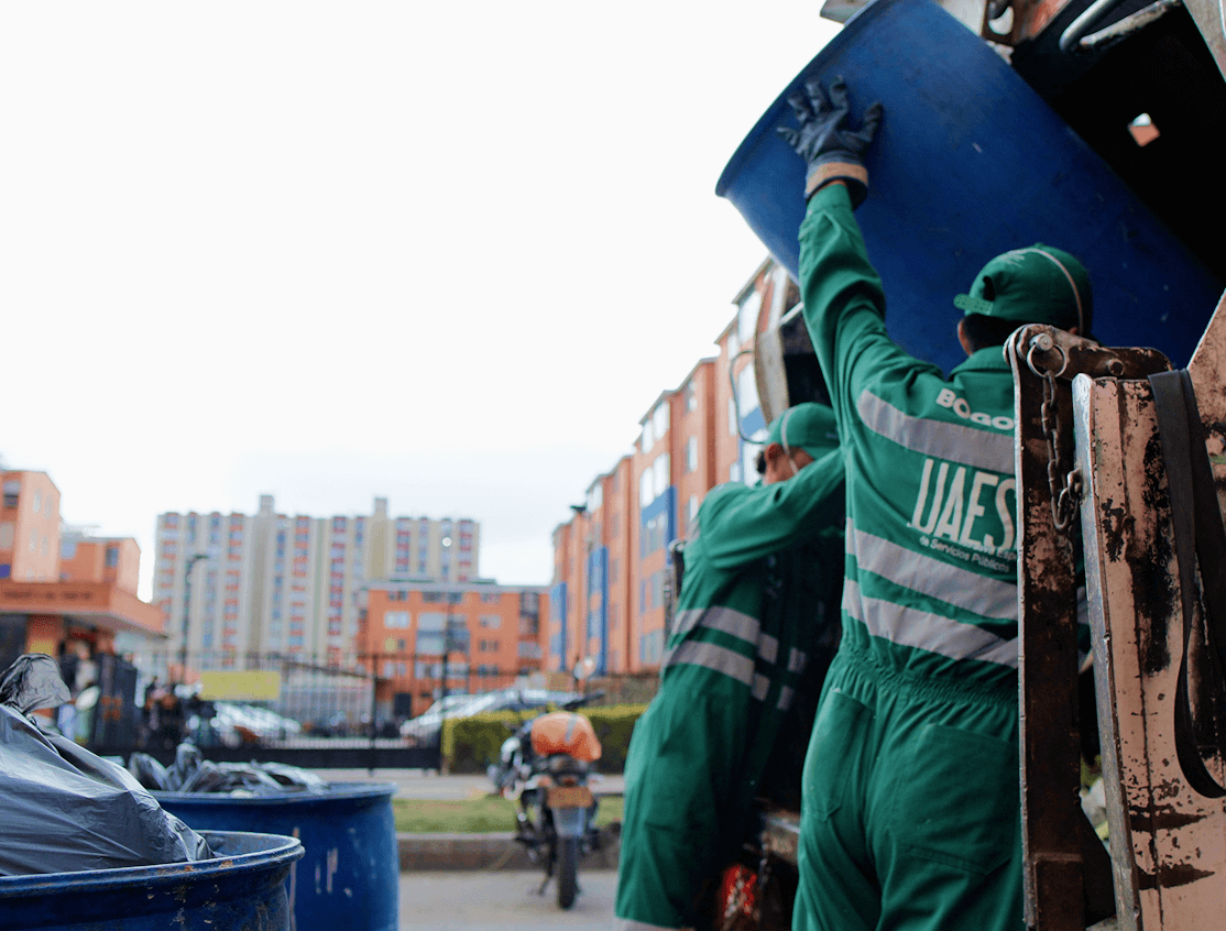 Dos colaboradores de Ciudad Limpia vacían un contenedor azul en un camión recolector durante una jornada de recolección de residuos en un entorno urbano residencial.