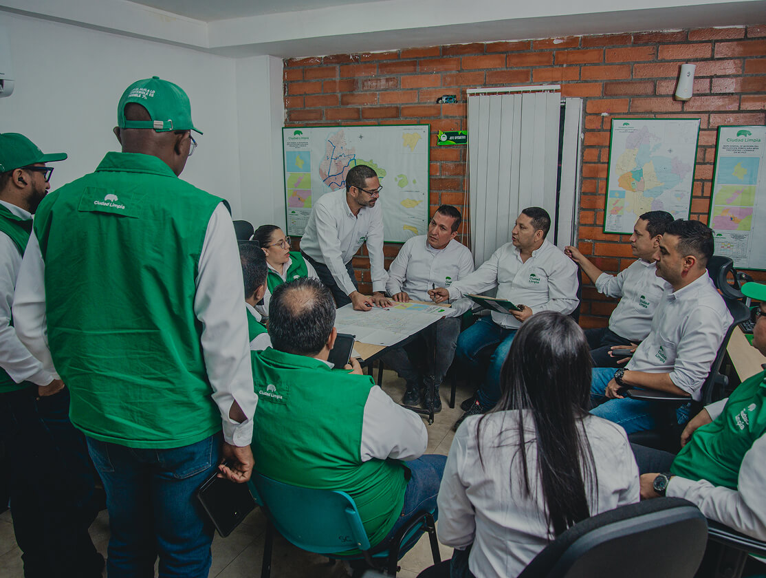 Fotografía de un grupo amplio de colaboradores de Ciudad Limpia reunidos en una oficina, ambientada con mapas de colores. Uno de ellos lidera la reunión desde una mesa con documentos. Los demás se encuentran sentados alrededor y cuentan con camisas manga larga color blanco y y chalecos verdes, con el logo de la empresa.