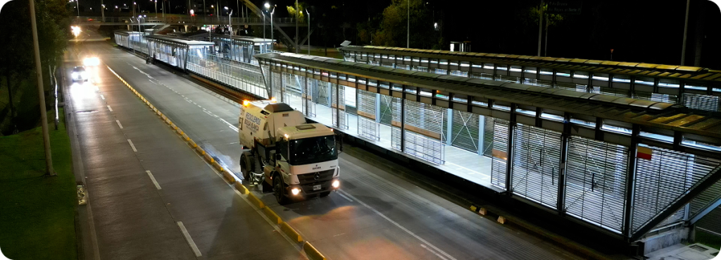 Barredora mecánica de Ciudad Limpia recorriendo una vía principal durante la noche, realizando labores de limpieza sobre el separador vial. A un costado se observan estaciones iluminadas de transporte público, vehículos en circulación y la infraestructura urbana bajo luz artificial.