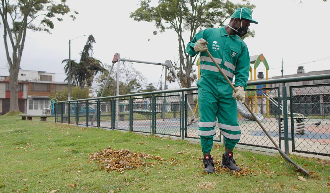 Fotografía de un colaborador de Ciudad Limpia portando un uniforme verde, guantes y tapabocas como protección, botas y una gorra. Se encuentra barriendo hojas secas en un parque con zonas verdes y áreas públicas, como césped, árboles y juegos infantiles.