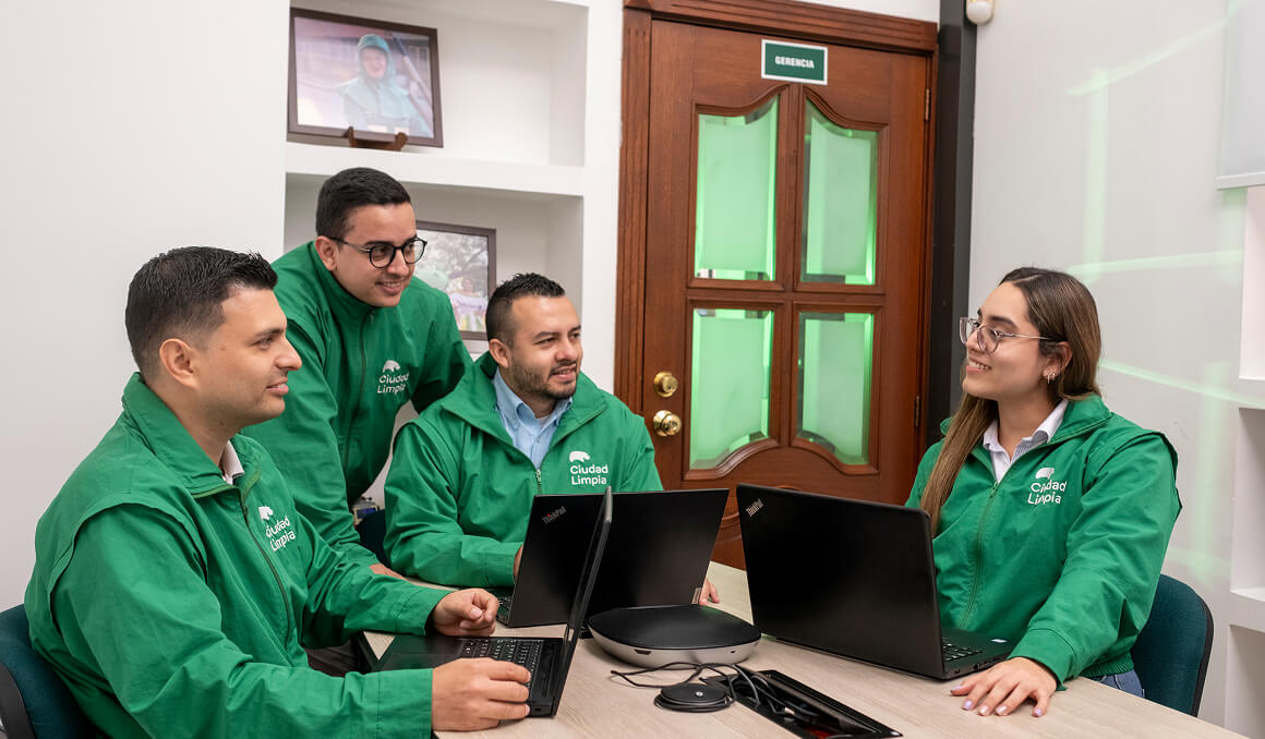 Foto de cuatro colaboradores de Ciudad Limpia reunidos en un entorno de empresarial. Todos lucen una chaqueta verde con el logo de la empresa y cuentan con computadores portátiles sobre una mesa amplia. En ella hay un micrófono para teleconferencias. Al fondo de aprecia la puerta de la oficina y algunas fotografías alusivas a la empresa, que adornan las paredes.