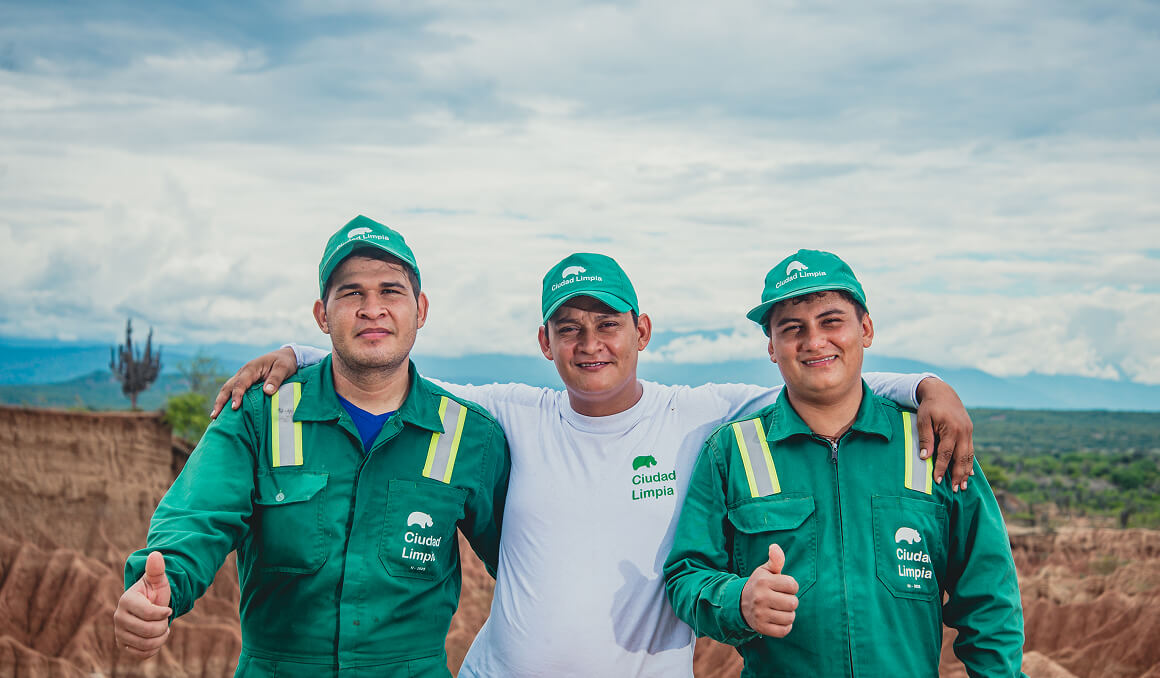 Fotografía de tres colaboradores de Ciudad Limpia con expresión sonriente. Los que están a los extremos izquierdo y derecho cuentan con un uniforme verde y gorra con el logo de la empresa. El de la mitad luce una camiseta manga larga blanca, también con el logo de Ciudad Limpia y una gorra verde. Al fondo se aprecia un paisaje colombiano con una franja terrosa, vegetación y un cielo azul acompañado con nubes blancas.