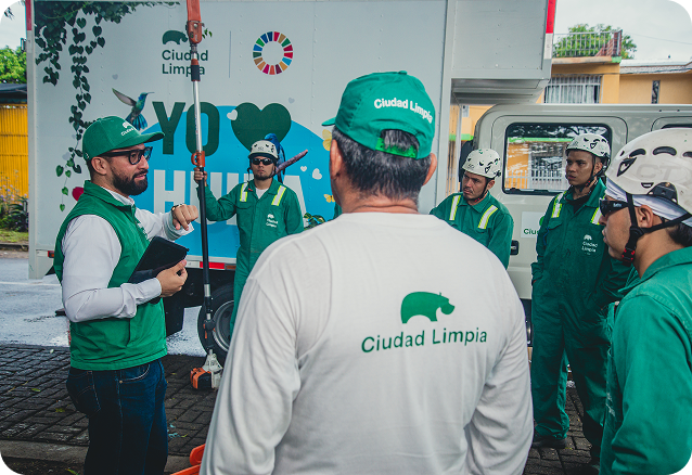 Fotografía de un equipo de colaboradores de Ciudad Limpia participando en una charla operativa al aire libre, junto a un vehículo institucional. Varios trabajadores con uniforme verde y cascos de seguridad escuchan indicaciones.