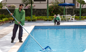 Colaborador de Ciudad Limpia realizando labores de mantenimiento y limpieza en una piscina, mientras otra persona trabaja en el área exterior del espacio.