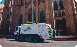 Camión recolector de Ciudad Limpia operando frente a una iglesia en un entorno urbano, mientras personal de la empresa realiza labores de recolección en el lugar.