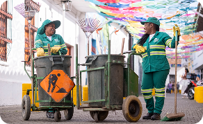Dos colaboradoras de Ciudad Limpia conversan y sonríen mientras realizan labores de limpieza en una calle, acompañadas de carros de recolección, escobas y elementos de señalización.